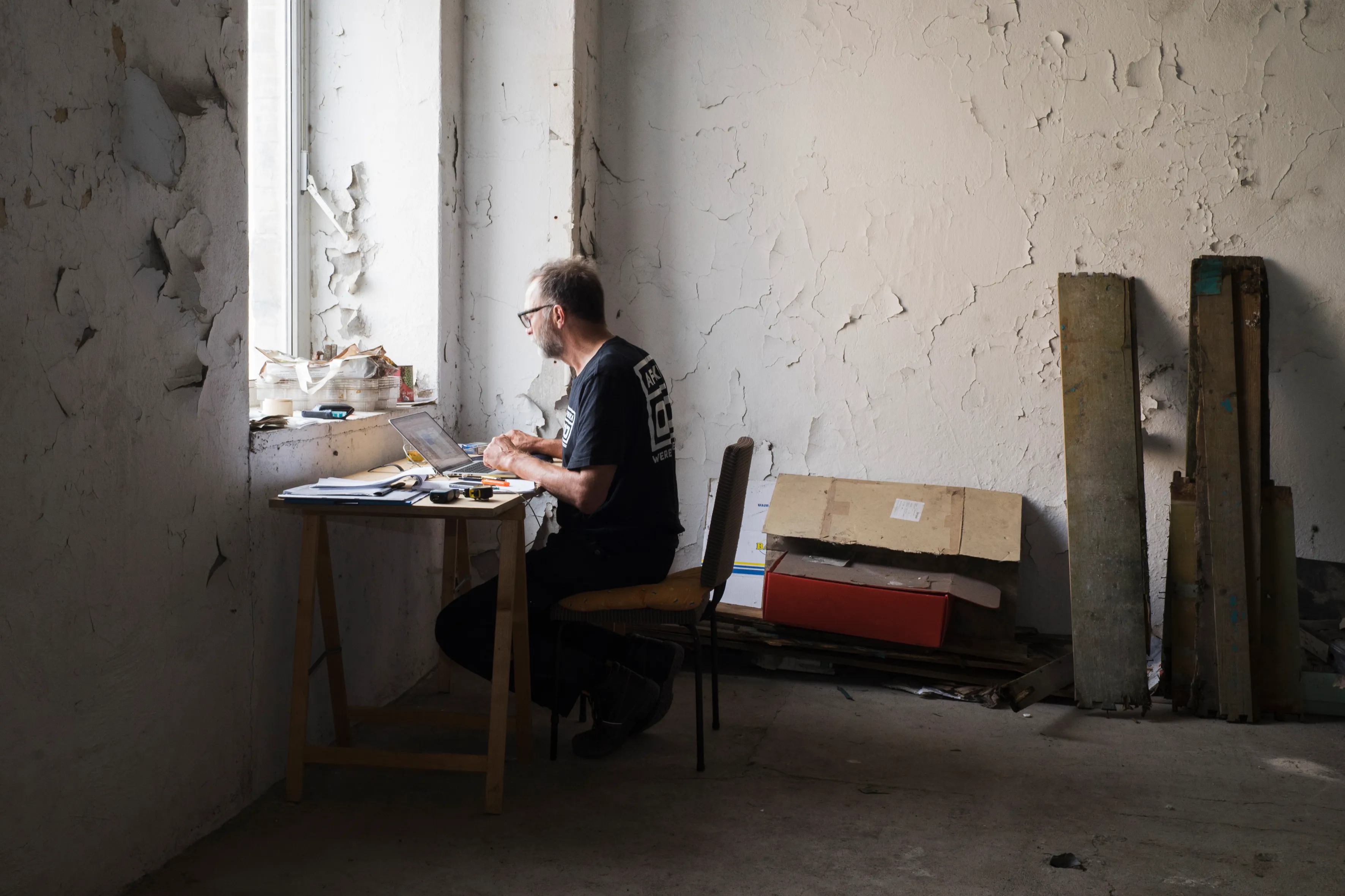 An artist working at a laptop by a window in a derelict room